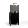 An olive green tote bag with black handles is shown hanging from the telescoping handle of a black hard-shell spinner suitcase on a clean white background.