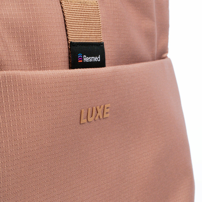 Close-up of a the Luxe tote dusty rose pink, showing the front pocket with the embossed brand name LUXE and a black Resmed logo tag on a beige webbing strap.
