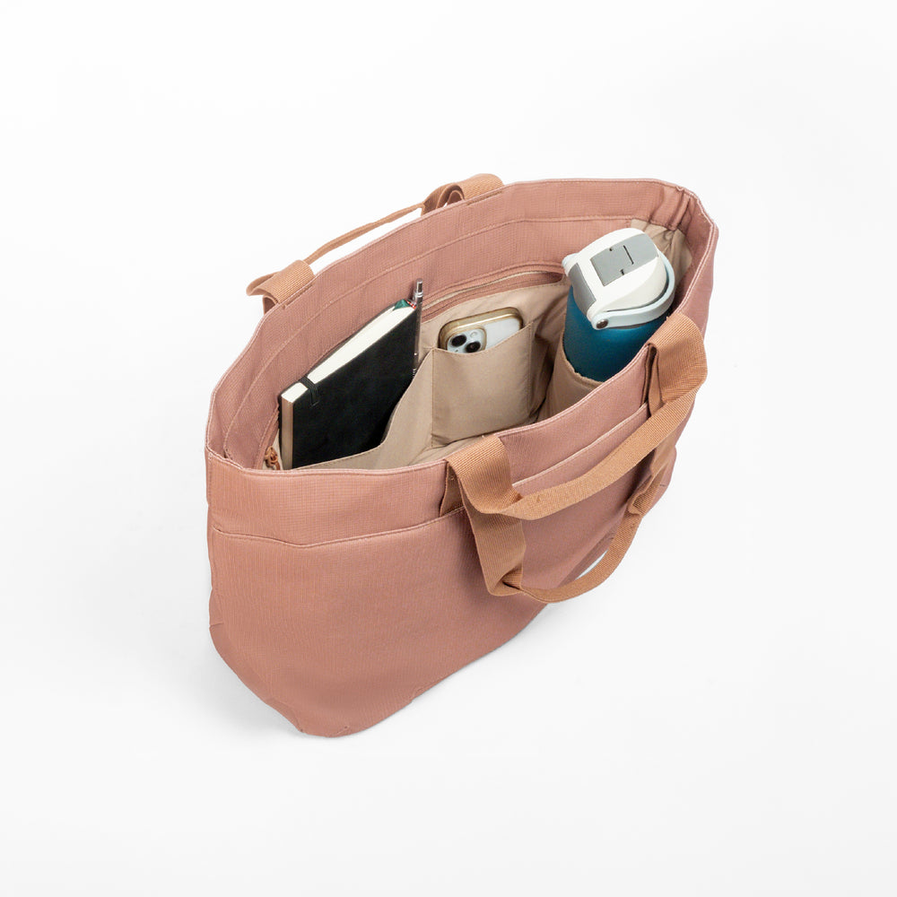 Luxe tote bag A dusty rose canvas tote bag shown open from an overhead angle, filled with a black notebook, a beige phone case, and a teal insulated tumbler, on a clean white background.
