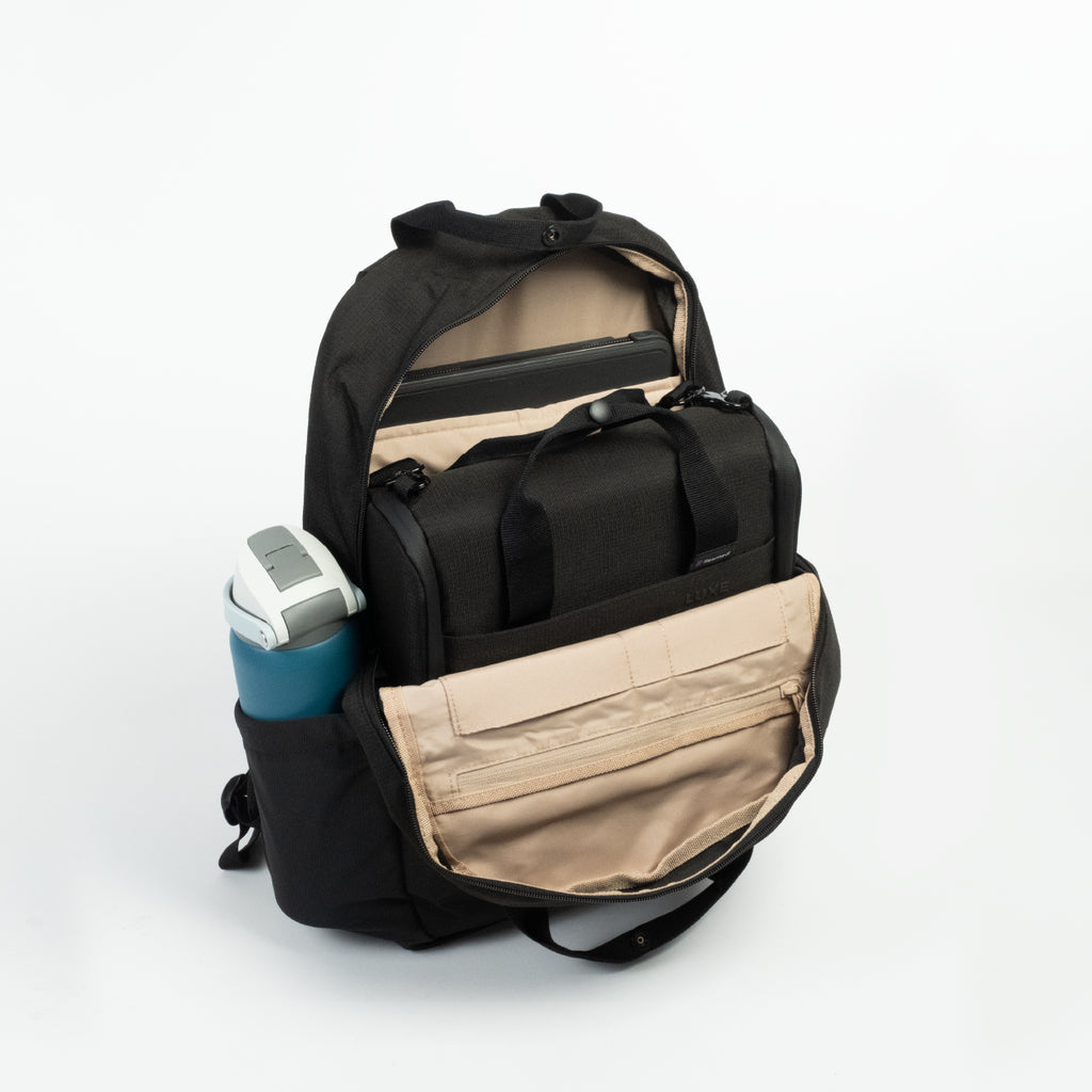 A black backpack shown open with a CPAP insert, on a white studio background, with beige interior lining and a side pocket holding a blue water bottle. 