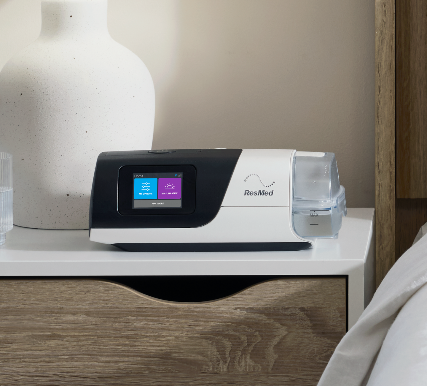 ResMed AirSense 11 AutoSet CPAP machine in black and white sitting on a wooden bedside table, with its colour touchscreen displaying 'My Options' and 'My Sleep View,' and an attached water chamber.